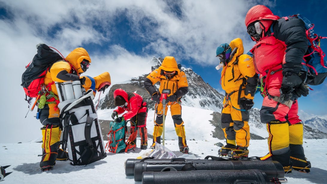 A team of six Everest expeditioners standing around a drill on South Col, dressed in protective geart