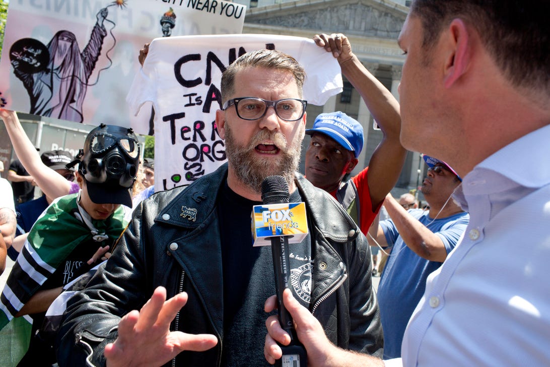 Gavin McInnes answers questions from a journalist in front of rally goers holding signs.