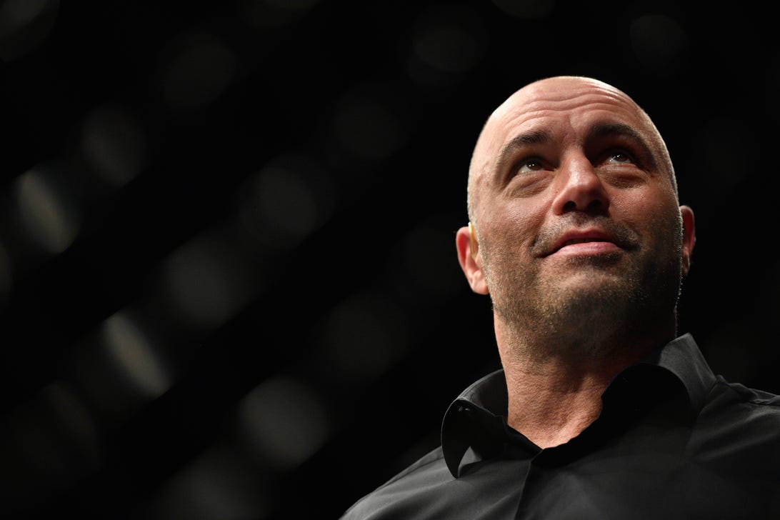 Joe Rogan, in a black button-up shirt, looks up with a smile while standing against a black background.