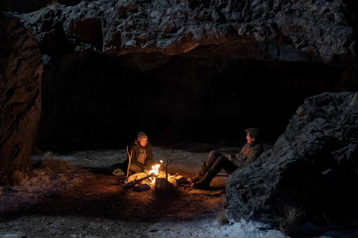 Ellie and Joel sit in front of a campfire among large rocks in The Last of Us