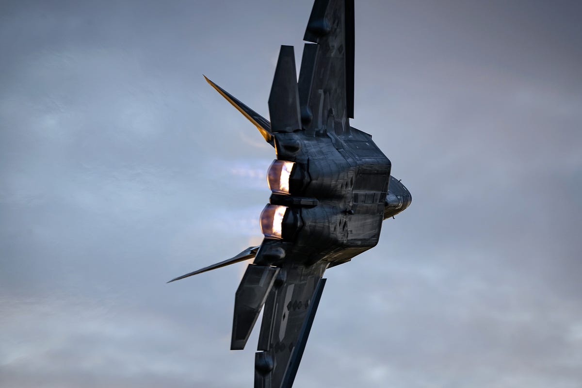An F-22 fighter, turned on its side, screams dramatically away from the camera into an overcast gray-blue sky, its jet engines lit by a fiery orange glow.