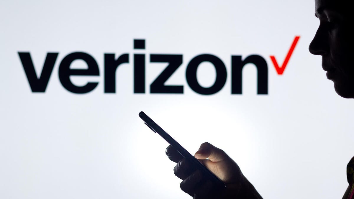 A person in silhouette holds a smartphone with the Verizon logo in the background