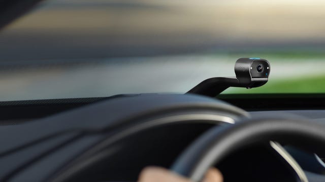 The Ring Car Cam, a dual-facing dashcam with Wi-Fi connectivity, sits atop the dashboard of a vehicle pulling past a bokeh background.