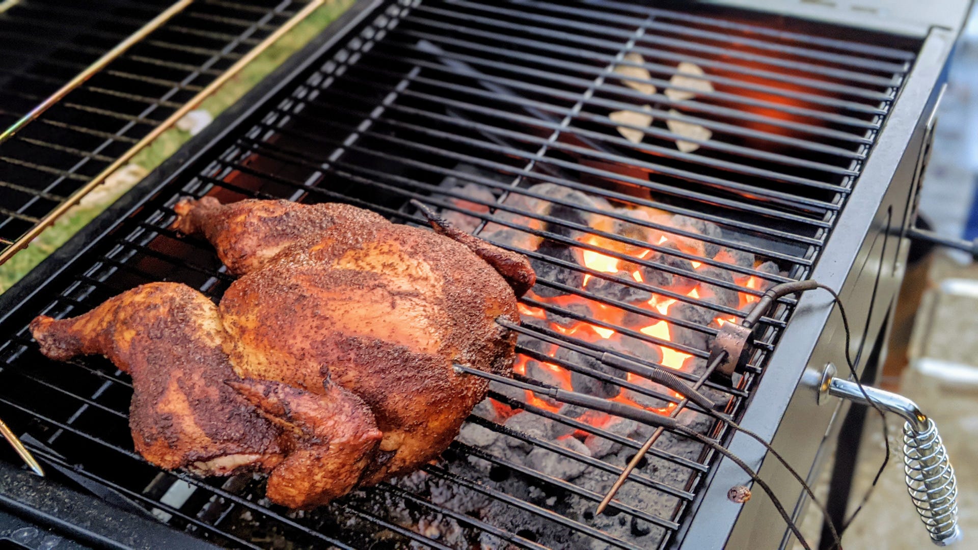 A butterflied chicken covered in barbecue rub with thermocouple probes in it, cooking on a rectangular grill