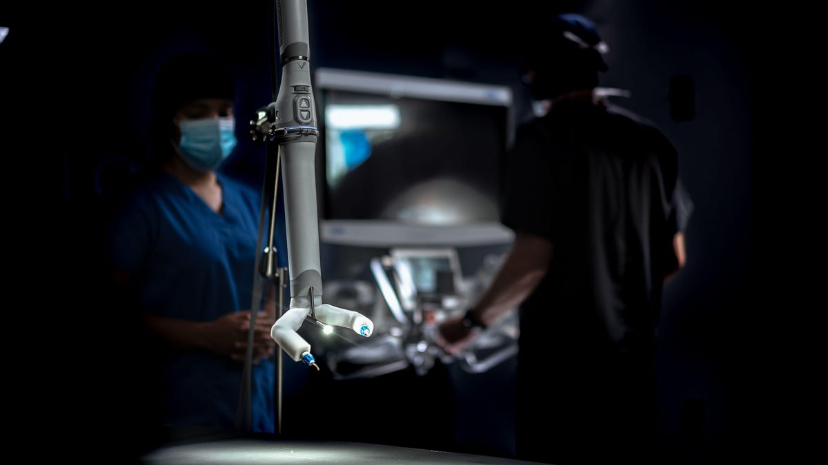 Doctors are seen in the background. In the foreground, the MIRA robot is lit up. It's a white rod with two appendage-like arms on one end, between which there is a tiny light fixture.