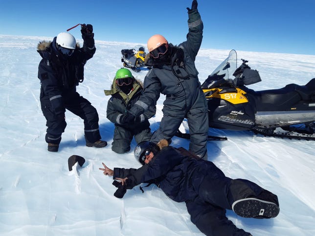 Four scientists pose against the Antarctic snow in enthusiastic positions around a dark meteorite. A snowmobile is off to the side.