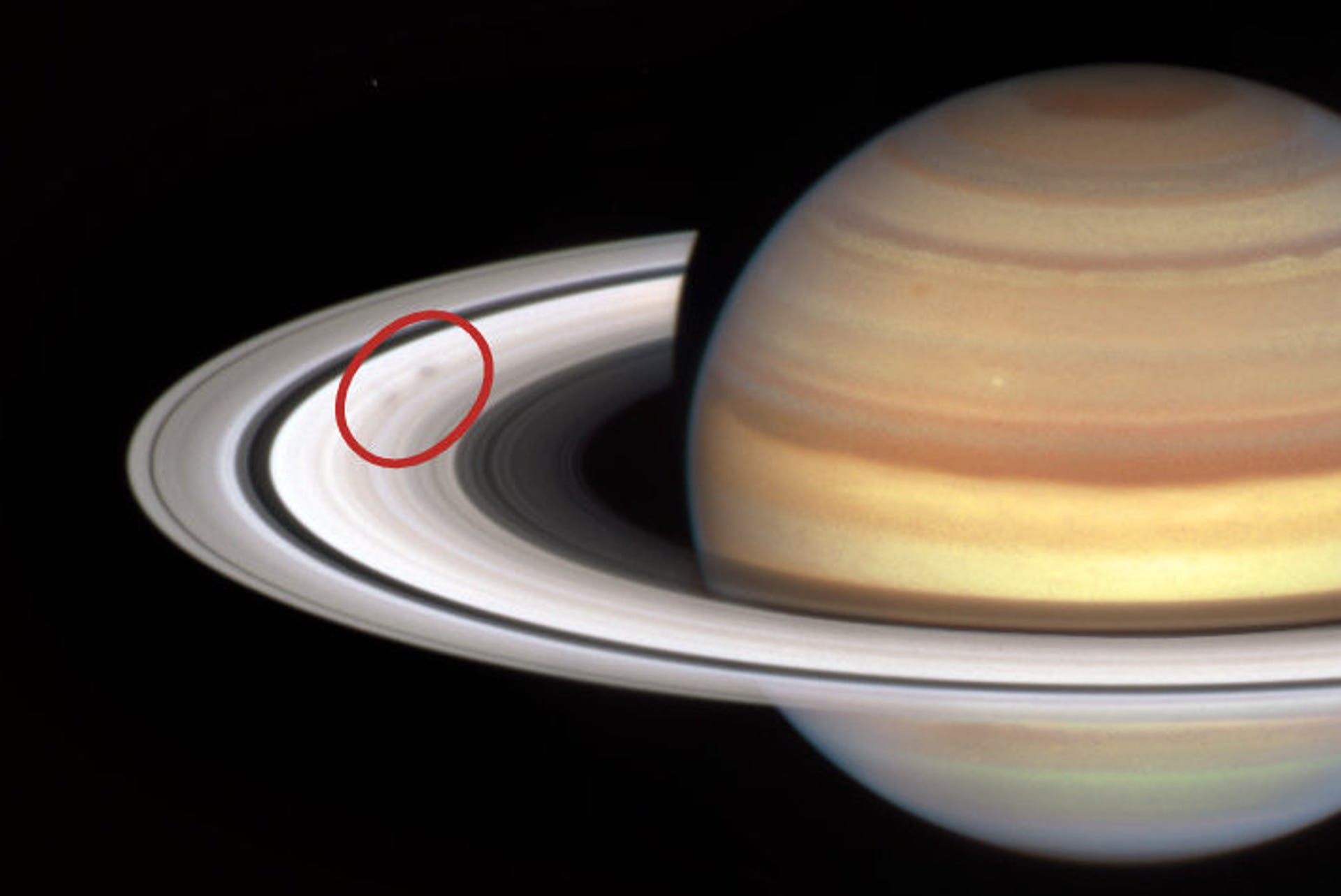 A close-up image of the planet Saturn. The rings are level with the viewer, and tilted slightly down. A red oval highlights the dark, smudgy 