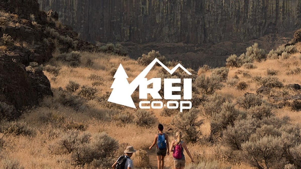 Three people hiking through the desert towards a large cliff face, with the REI Co-op logo superimposed.
