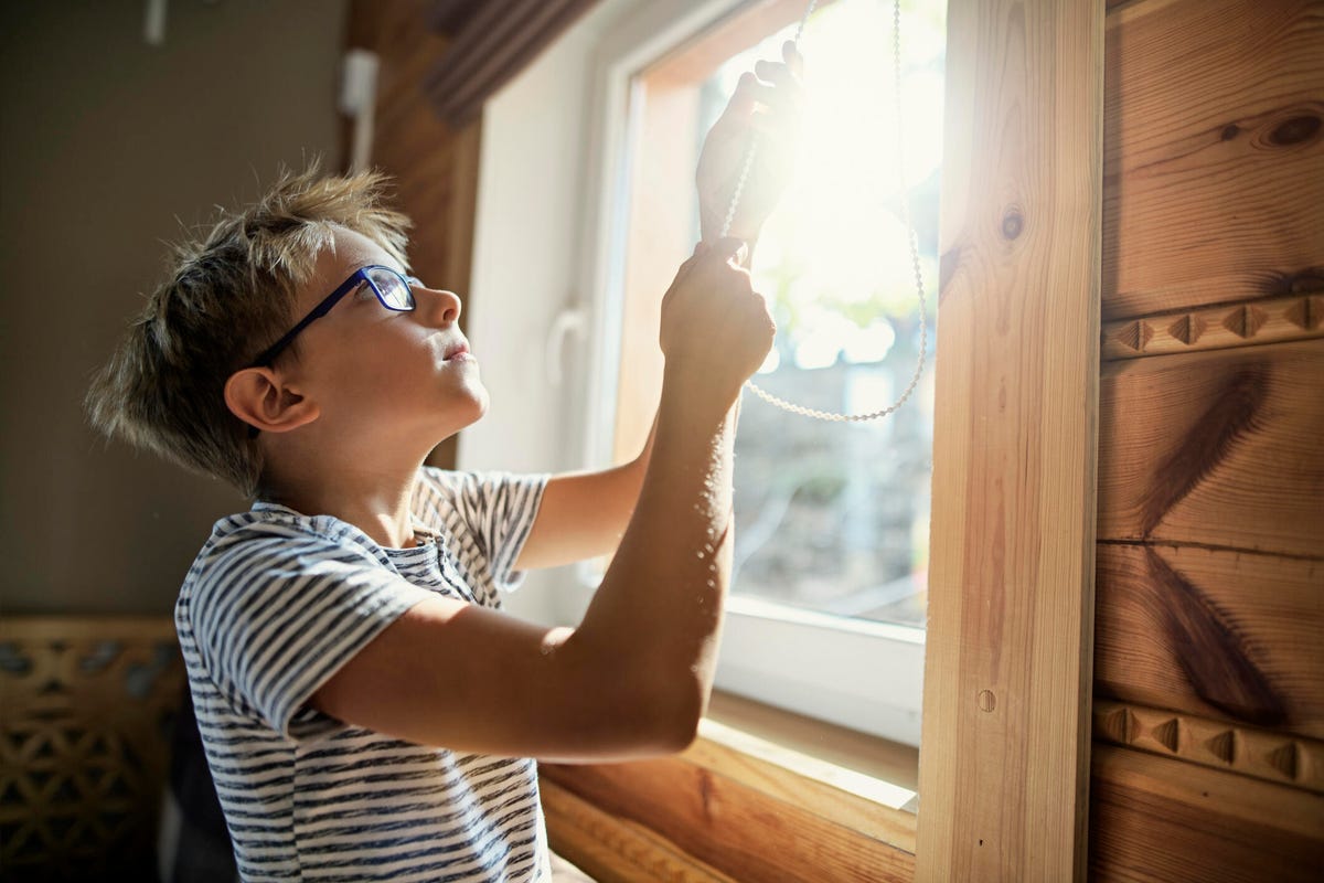 A boy adjusts window blinds on a sunny day.