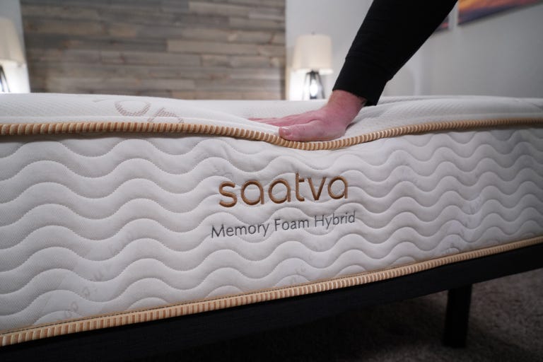 The Saatva Memory Foam Hybrid mattress logo being pushed down on by a hand in a moody-lit room.