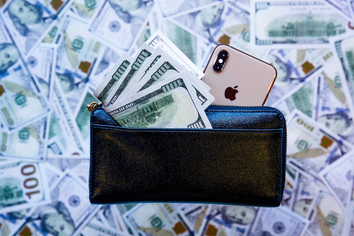 an iPhone peeking out from behind a wallet overflowing with hundred dollar bills