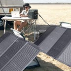 Man in hat using his laptop with solar panels laid out on the beach