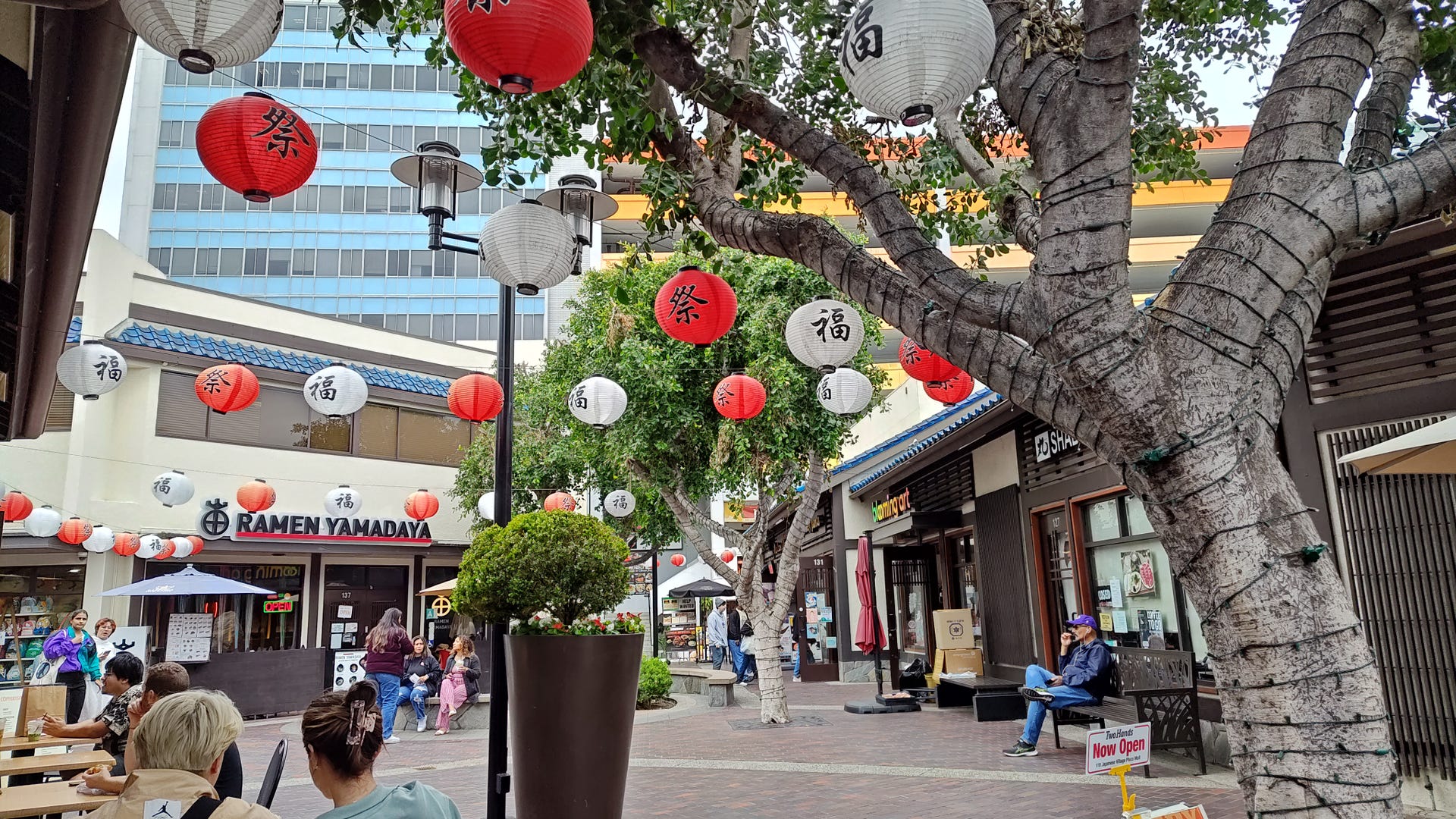 A scene in the Little Tokyo neighborhood of Los Angeles, where red and white lanterns strung between trees and shops hang over passersby.