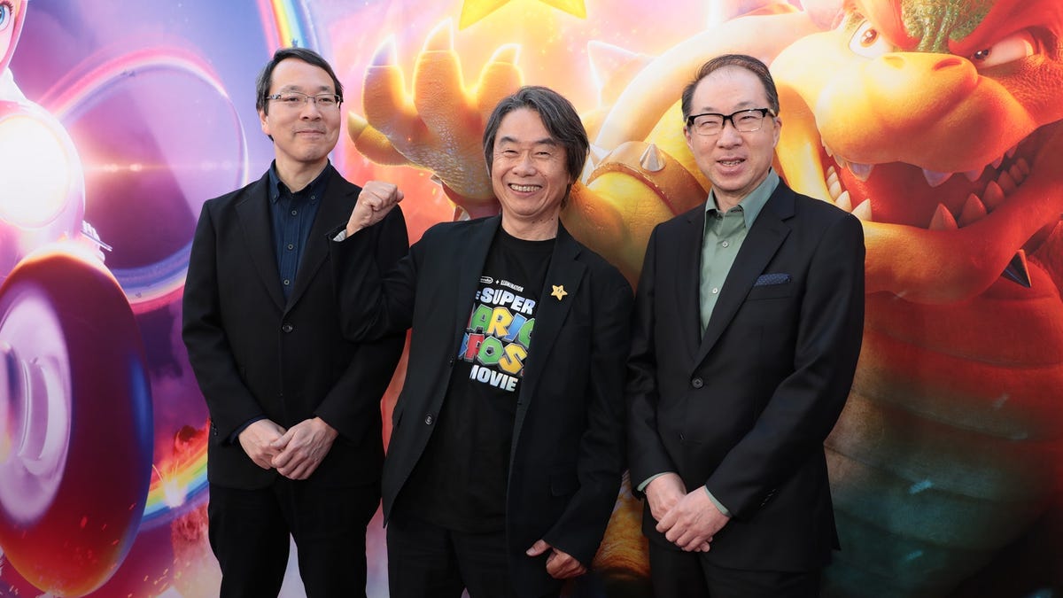 Three men jovially pose on a red carpet in front of a banner showing Bowser, Mario and other characters from the film.