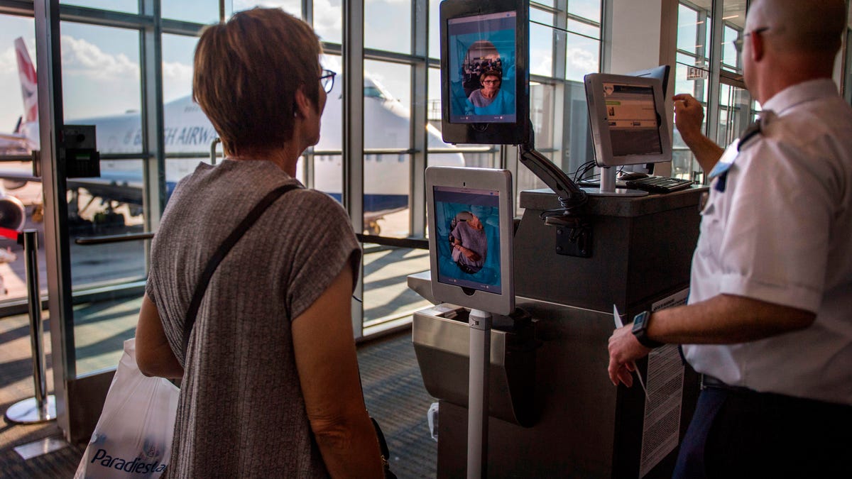 A woman stands in an airport boarding gate in front of a kiosk with two screens showing a photo of her face.