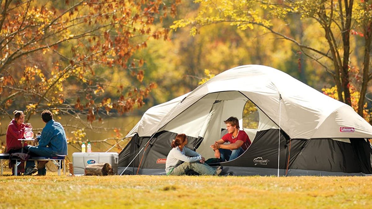 A man sits in a Coleman camping tend, speaking to a woman sitting outside of the tent, while another man and woman sit and talk near a cooler.