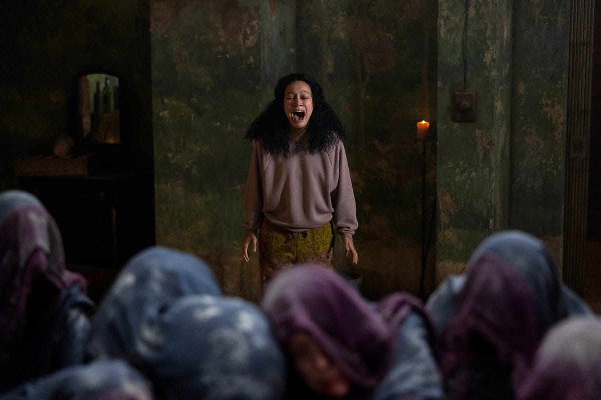 A girl standing behind hooded figures screaming in a basement