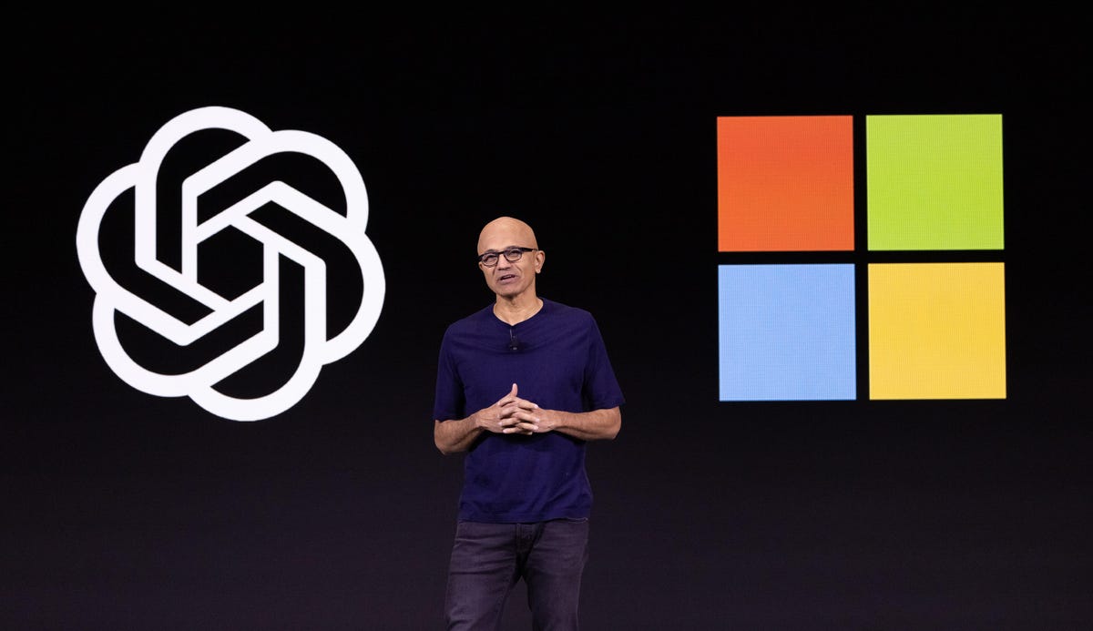 Microsoft CEO Satya Nadella speaking while standing between logos for OpenAI and Microsoft
