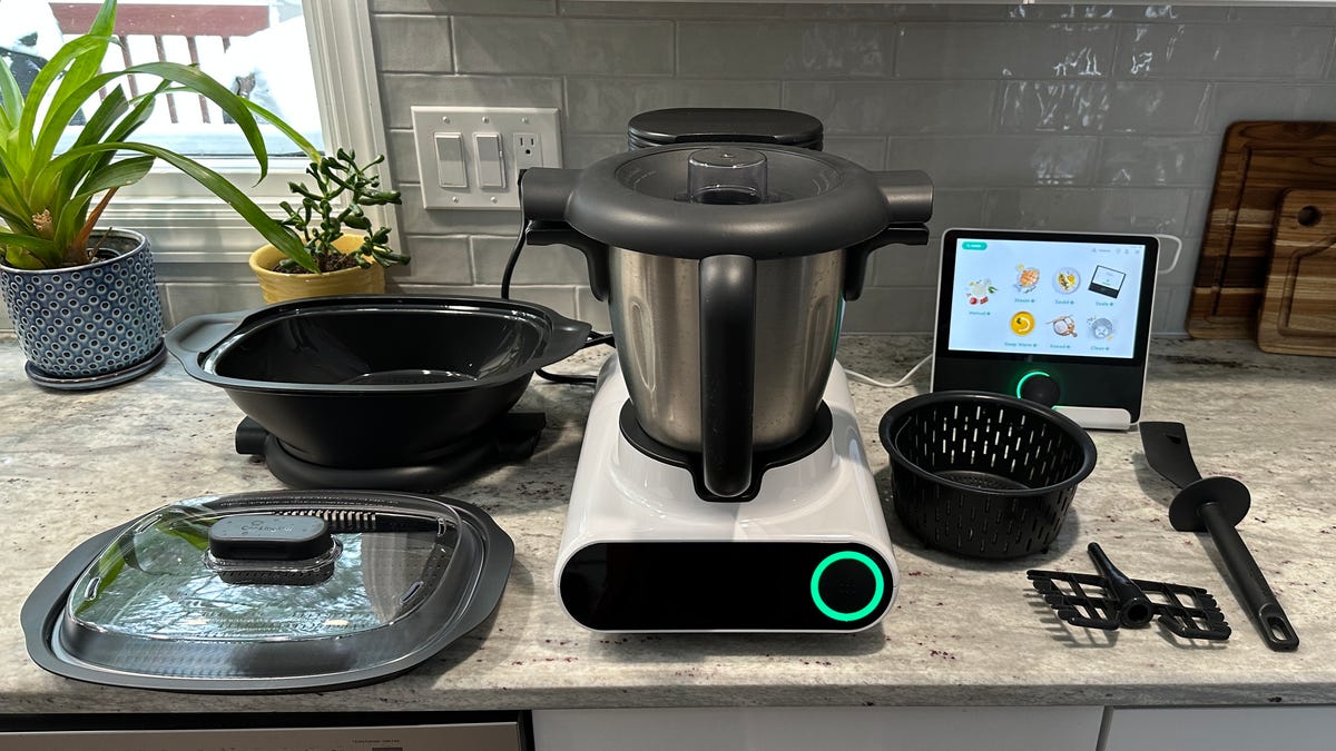 The CookingPal Multo on a kitchen counter with all of the included accessories: Smart Kitchen hub, simmer basket, spatula, butterfly whisk, and 3-piece steamer set