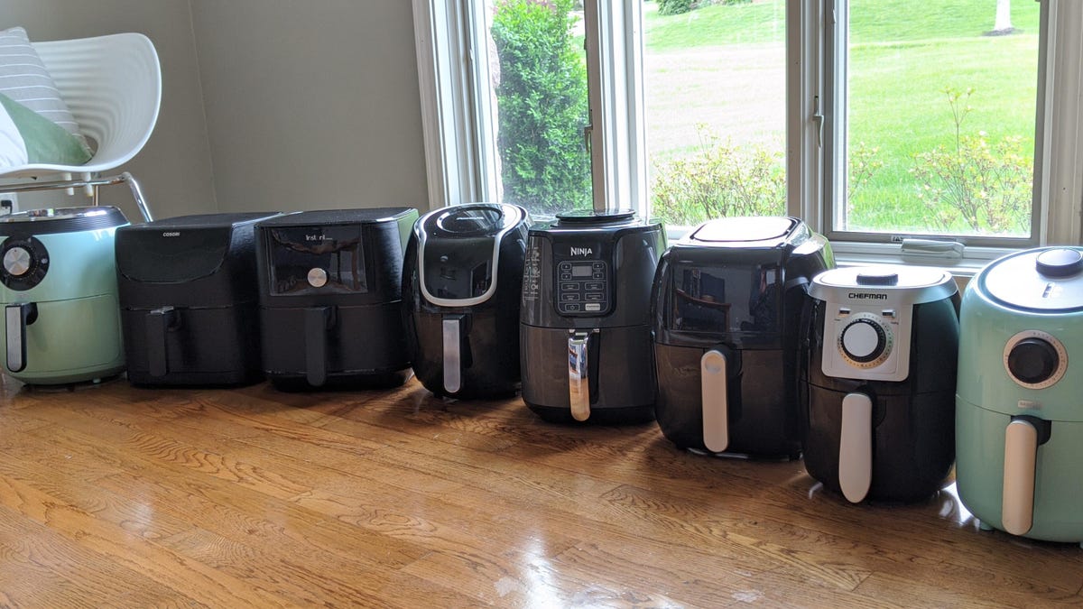 Eight air fryers lined up on a counter in front of a window