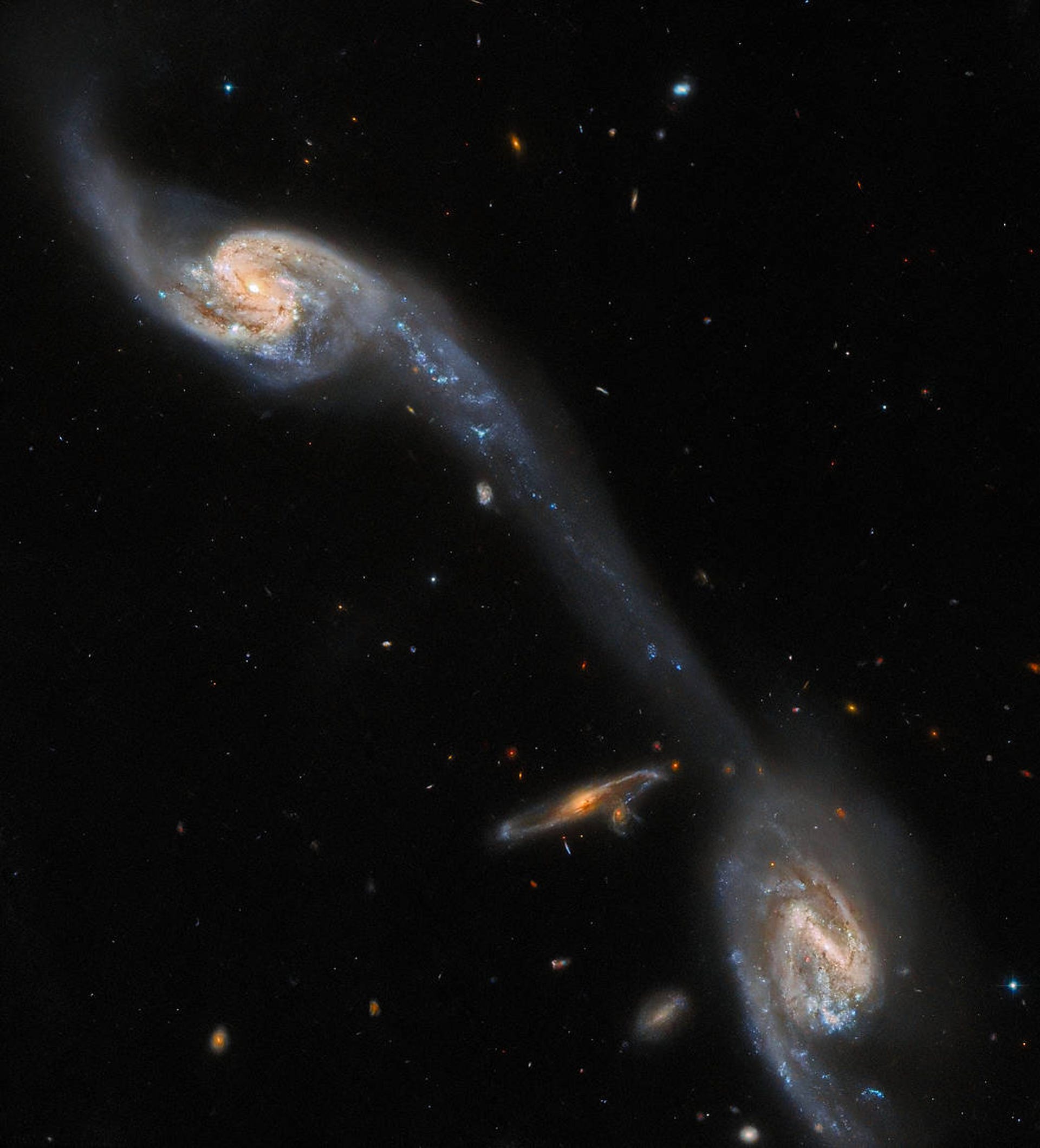 Two galaxies in the foreground appear connected by a wispy, luminous bridge of interstellar dust. A third galaxy is seen in the background, surrounded by many tiny specks of light that represent other, more distant, galaxies across the universe.