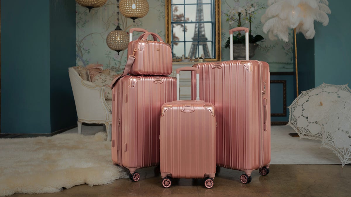 A 4-piece set of peachy-pearlescent luggage sits in the middle of a furnished room with a view of the Eiffel Tower.