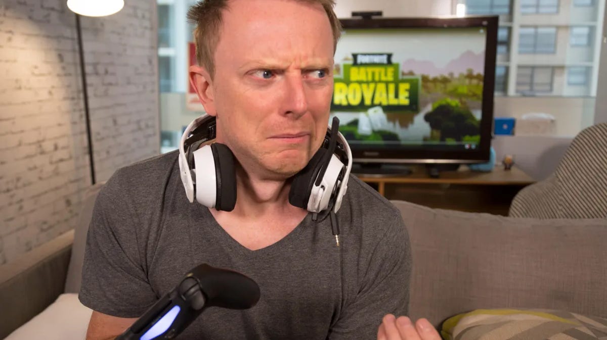 Mark Serrels grimacing, headphones around his neck and a TV screen behind him with the words Battle Royale
