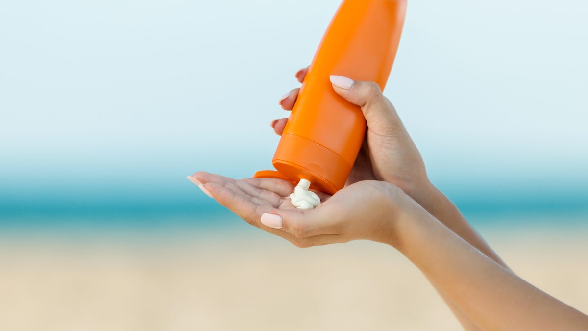 Person pouring sunscreen into hand with a beach in the background