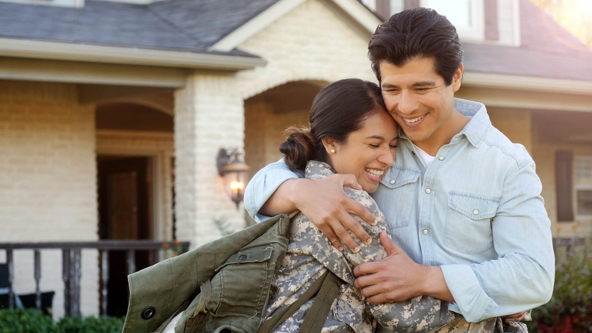 A woman in military attire hugging a man in front a white brick house.