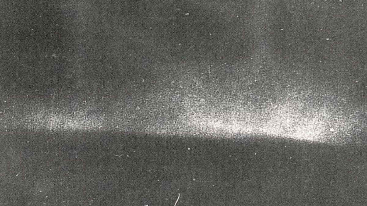 Ghostly black and white view shows curve of horizon with lighter splashes above it, indicating the Northern Lights.