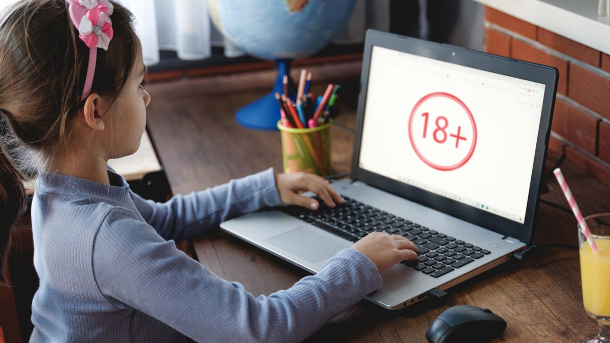A young girl sits at a laptop, whose screen shows an image reading "18+"
