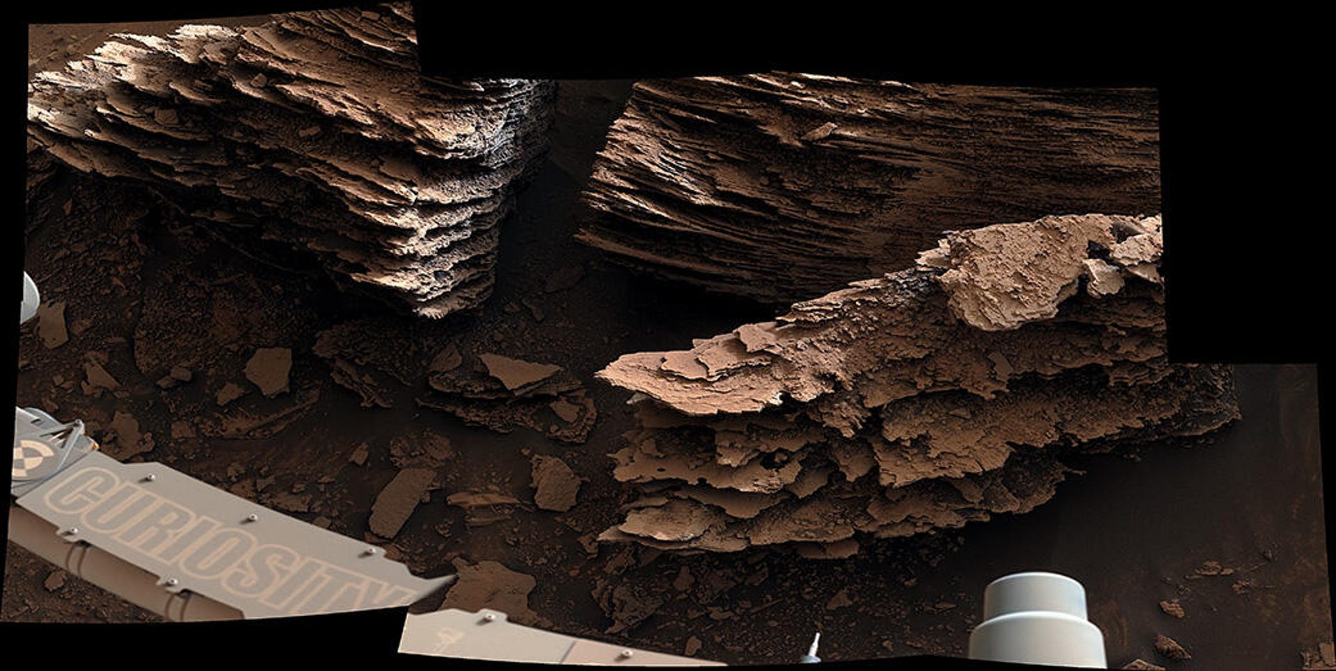 A part of the Curiosity rover appears in the corner of a close-up showing a finely layered, crispy-looking rock formation.