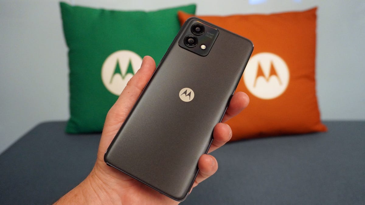 A black phone with a Motorola &apos;M&apos; logo is held in hand in front of two pillows, also bearing the Motorola logo.