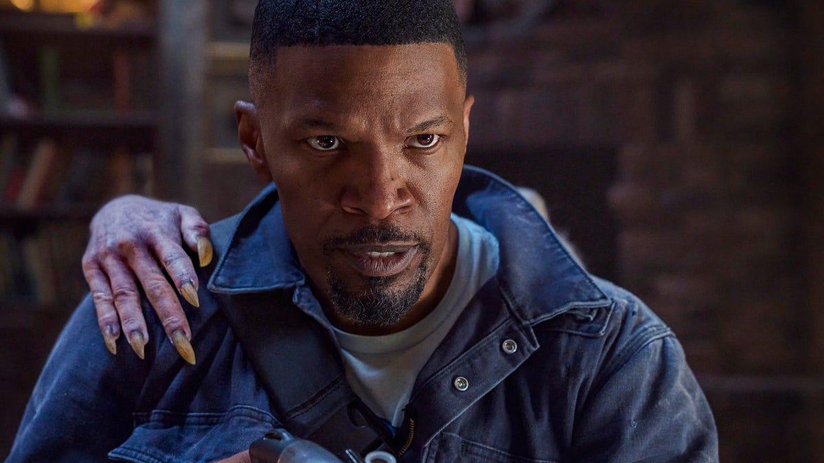 Jamie Foxx holding a gun, with a long-fingernailed hand touching his shoulder from behind