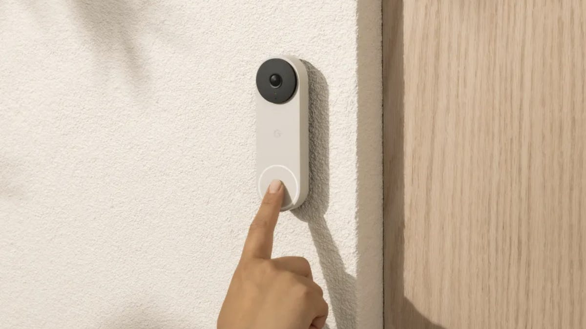 A finger presses the Nest Video Doorbell on white stucco beside a wood door.