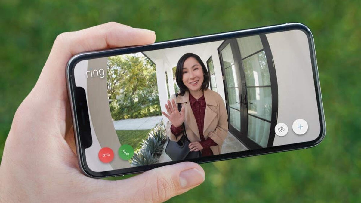 A smartphone in someone&apos;s hand showing a landscape view of the Ring Doorbell live view with a woman waving on the screen.