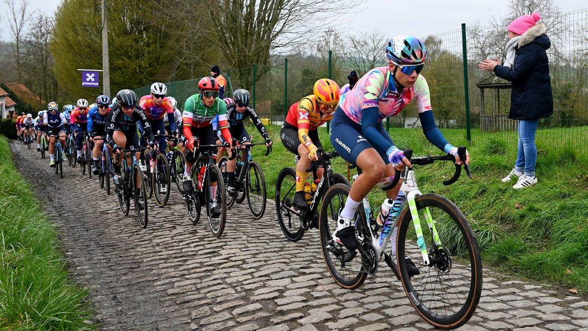A large group of competing riders in the Tour of Flanders passing along a cobbled street while a spectator claps by the roadside.