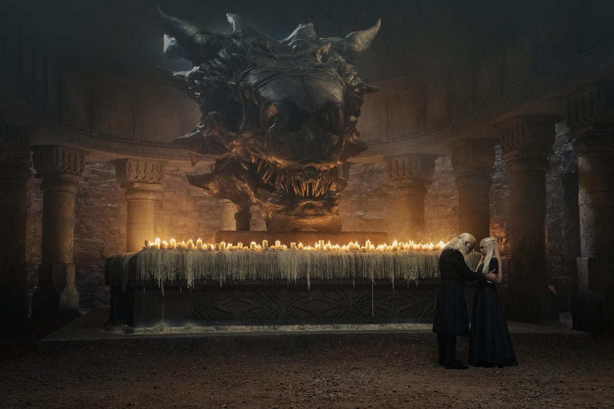 Viserys and Rhaenyra in a tete-a-tete in front of a long altar covered with lit candles, under a large stone dragon's head