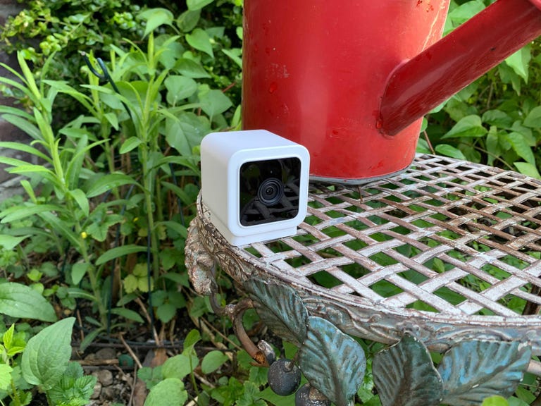 A small Wyze camera on outside plant stand next to a red watering can