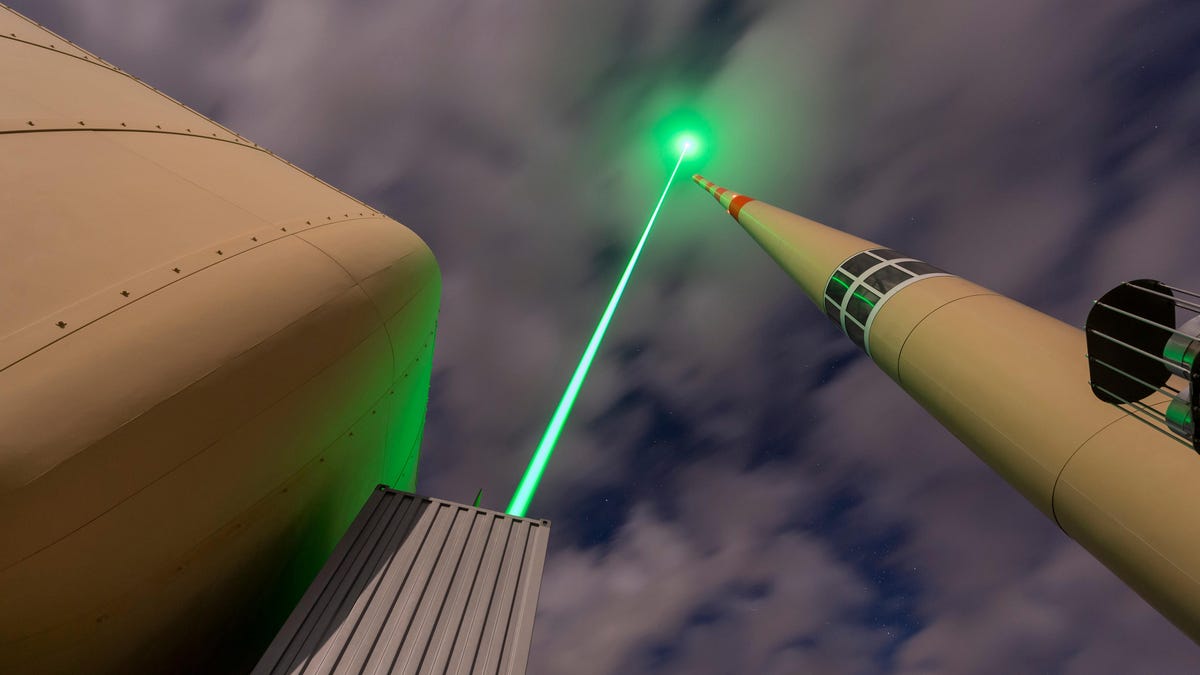 A green laser is projected from a large grey box into a cloudy sky. On its right is another tower and its left, a large white tank