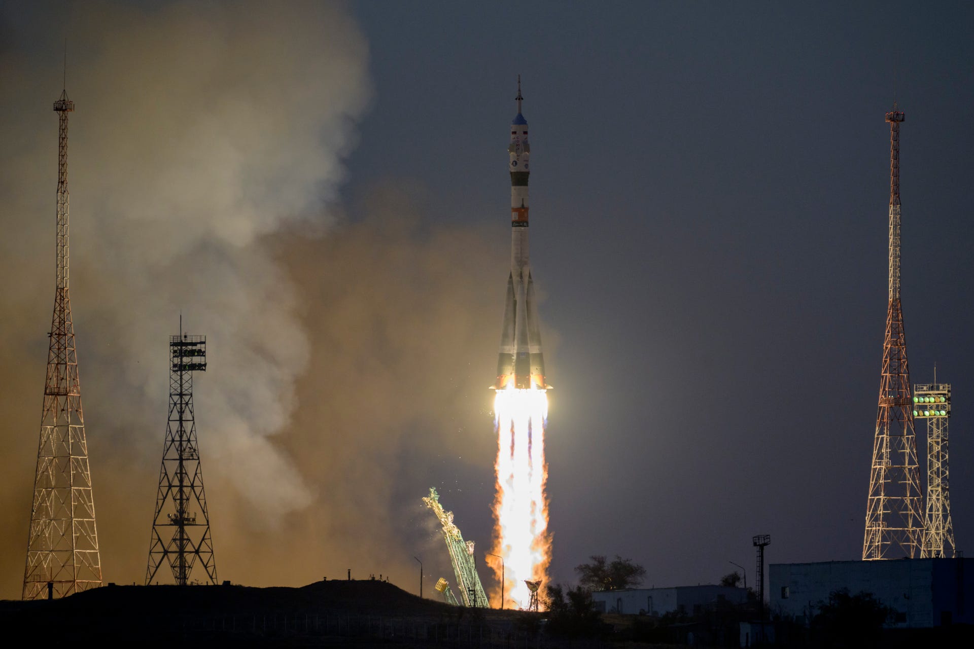 Russian rocket blasts off in a fiery blaze with launch site equipment towers nearby.