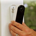 A hand places the Arlo 2K doorbell on its base, attached to white external house trim.