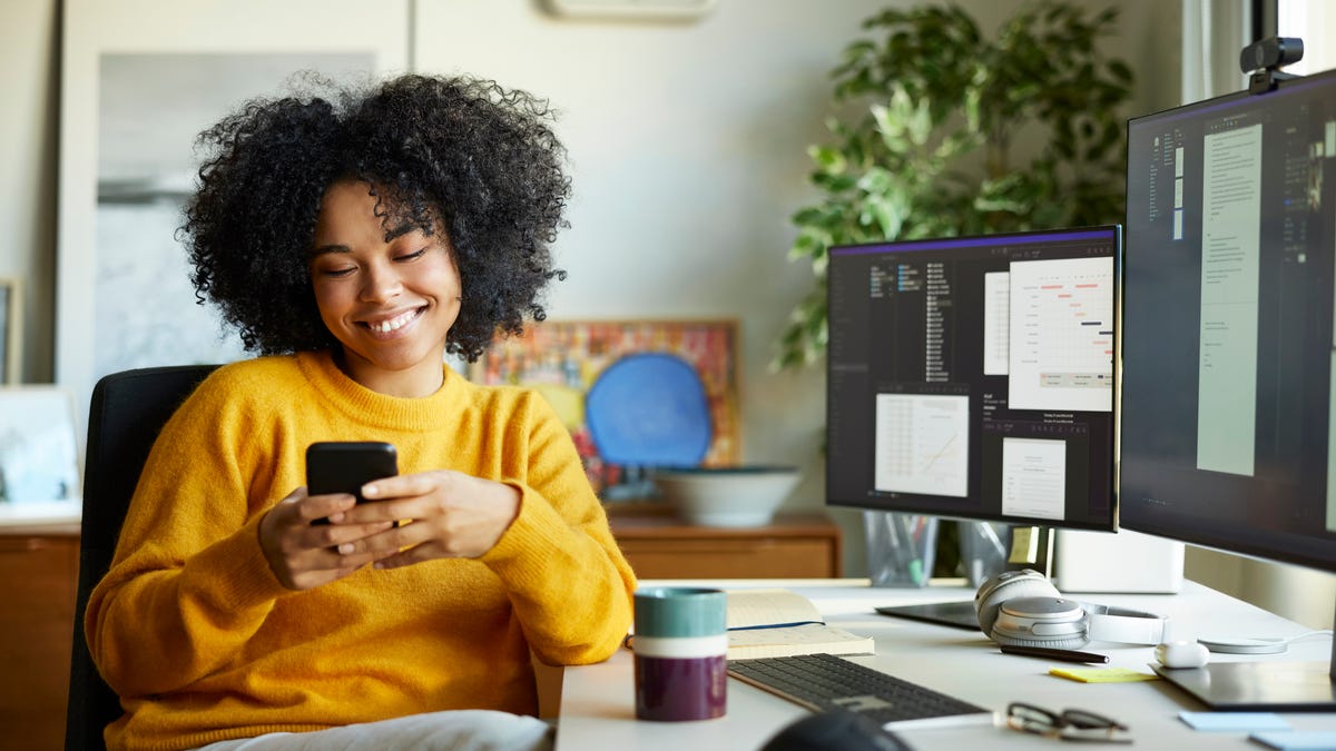 Woman at a desk smiling at a smartphone she is holding in her hands