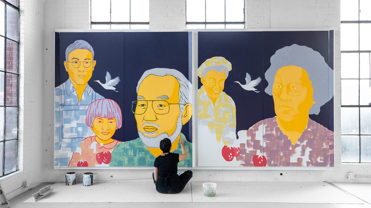 A woman sits cross-legged on the floor and paints a wall-sized mural of the faces of Japanese Canadian farmers.