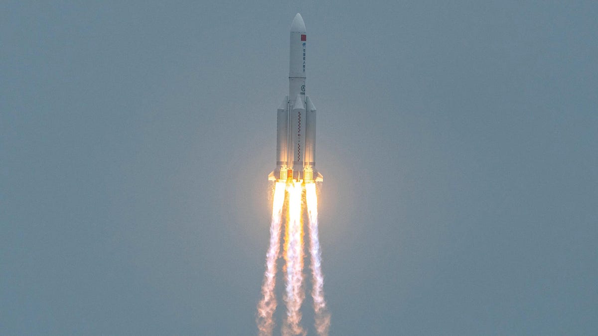 A white rocket, with 3 flames emanating out of the bottom, ascending against a grey cloudless sky