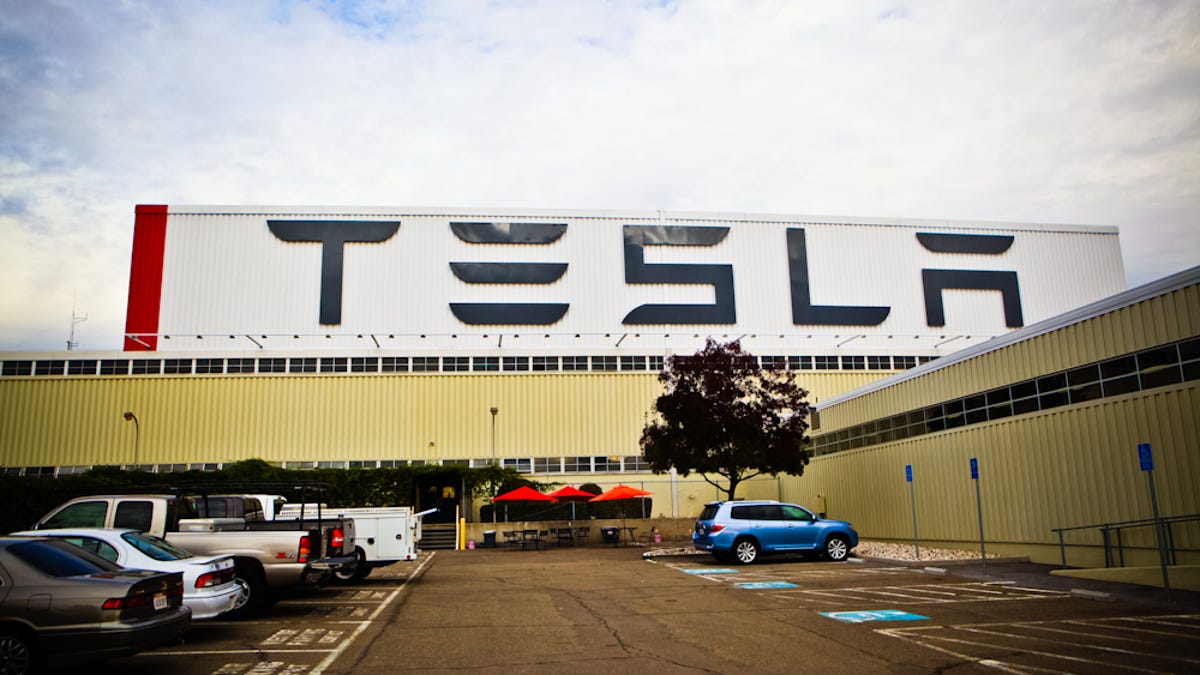 A large warehouse with TESLA on the sign, with parked cars in front.