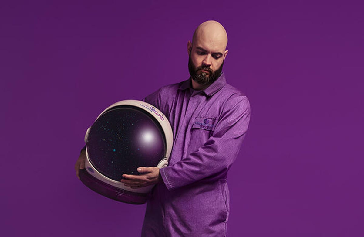 Man dressed in purple jumpsuit holding a space helmet