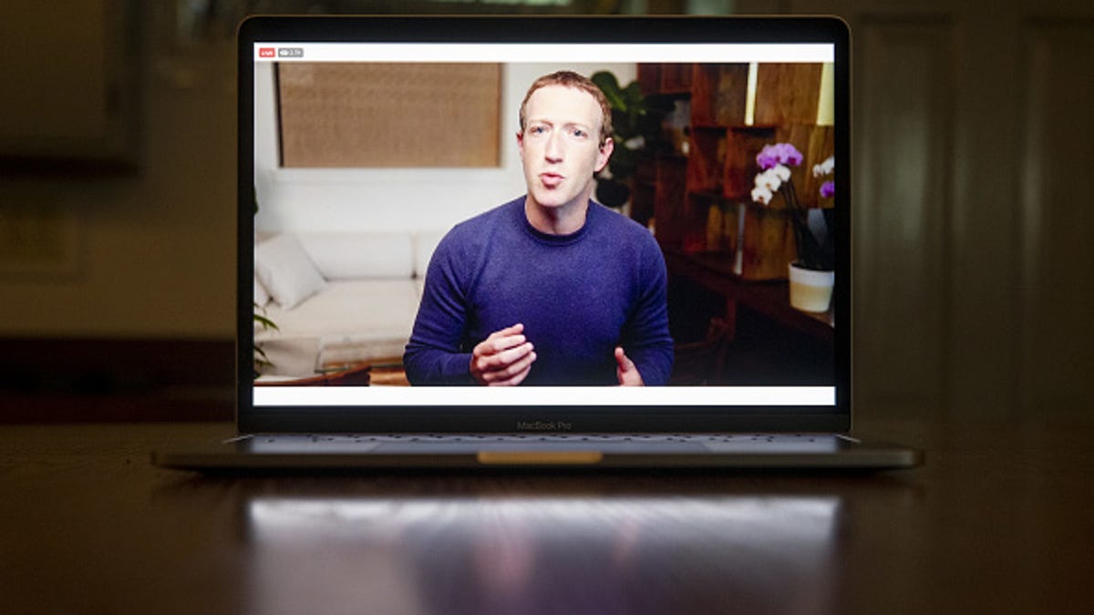 Mark Zuckerberg on a laptop screen, speaking from a room with a couch and wooden shelving with flowers
