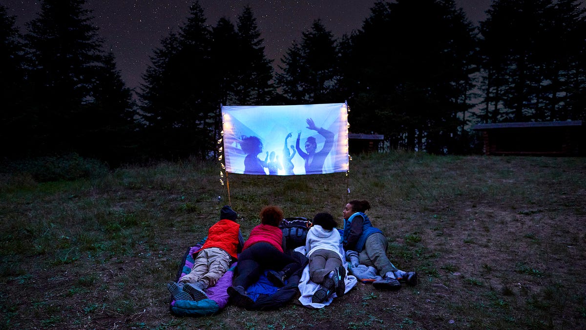 استلقى أربعة أطفال على العشب تحت النجوم يشاهدون فيلمًا على شاشة صغيرة.