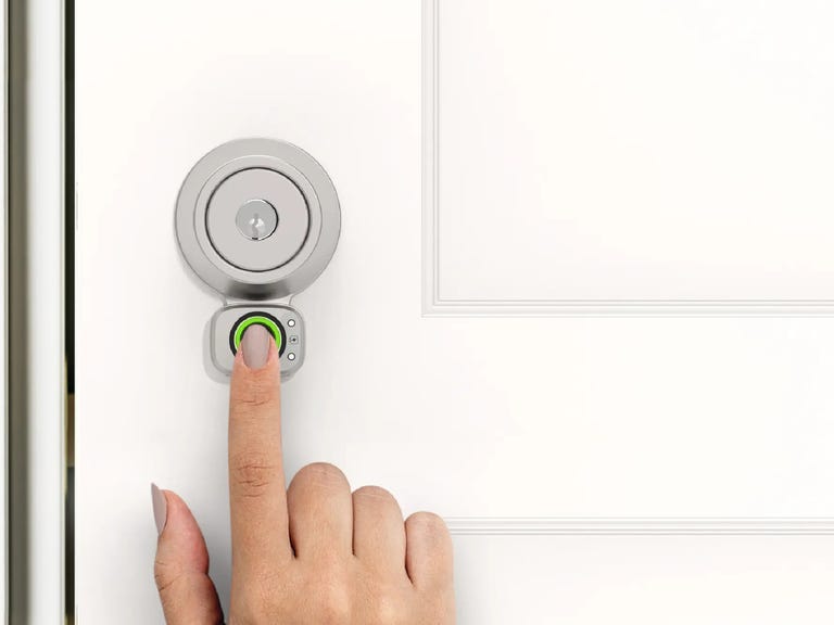 A finger touches the Lockly fingerprint reader installed on a metal deadbolt on a white door.